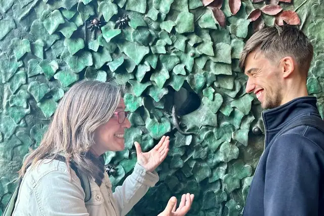 Two people facing each other, smiling and gesturing, in front of a wall covered in green, leaf-like sculptures.